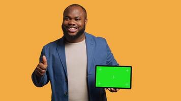 Jolly man holding isolated screen tablet, doing recommendation. Cheerful BIPOC person pointing towards chroma key device, doing thumbs up sign, studio background, camera A video