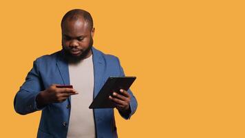 Smiling man scrolling on tablet touchscreen, using internet and credit card to pay for products. African american person happily browsing online shopping websites, studio background, camera A video