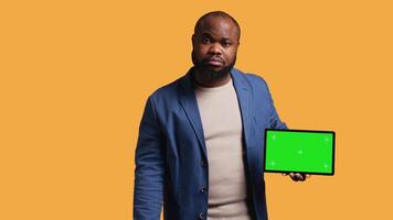 Upset african american man shows thumbs down sign gesturing holding isolated screen tablet, disapproving. BIPOC person does rejection hand gesture regarding mockup device, studio background, camera B video