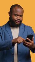 Vertical Portrait of energetic happy man holding smartphone celebrating, throwing fists in air from excitement. Overjoyed BIPOC person reading good news on phone, cheering, studio background, camera A video