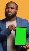 Vertical Portrait of african american man doing influencer marketing using green screen tablet, studio background. Smiling BIPOC person holding empty copy space mockup device, camera A video