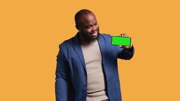 Portrait of man doing influencer marketing using isolated screen phone, showing thumbs up sign, studio background. Merry BIPOC person holding empty copy space mockup cellphone, camera B video