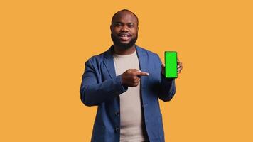 Smiling african american man presenting cellphone with green screen display, isolated over studio background. Cheerful BIPOC person creating promotion with blank copy space mockup phone, camera B video