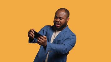 African american man playing games on cellphone in landscape mode, enjoying leisure time. Gamer enjoying game on mobile phone, having fun, studio background, camera A video