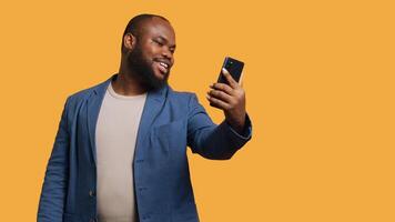 africain américain narcissique homme en utilisant téléphone portable à prendre selfies de tout angles. vaine social médias utilisateur prise Photos en utilisant téléphone selfie caméra, souriant Heureusement, studio arrière-plan, caméra b video