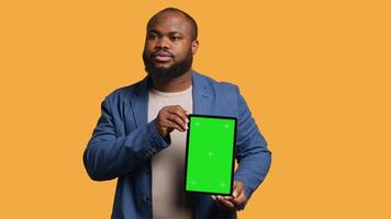 Portrait of african american man doing influencer marketing using green screen tablet, studio background. Smiling BIPOC person holding empty copy space mockup device, camera A video