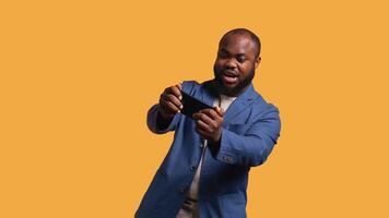 African american man playing games on cellphone in landscape mode, enjoying leisure time. Gamer enjoying game on mobile phone, having fun, studio background, camera B video