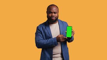 Upset african american man showing thumbs down sign gesturing holding green screen phone, disapproving. BIPOC person doing rejection hand gesture regarding smartphone, studio background, camera B video