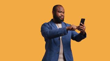 Smiling african american man greeting friends during teleconference meeting using smartphone, studio background. Friendly person waving hand, saluting mate during internet call, camera B video