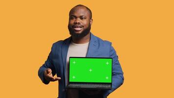 Smiling man holding isolated screen laptop, doing recommendation, talking with viewers. Cheerful BIPOC person pointing towards chroma key notebook, studio background, camera A video