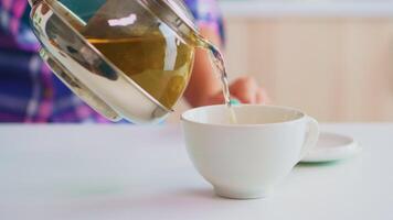 vert thé écoulement de théière dans lent mouvement. proche en haut de thé de le bouilloire lentement verser dans porcelaine tasse dans le cuisine dans le Matin à petit-déjeuner, en utilisant tasse à thé et sain à base de plantes feuilles. video