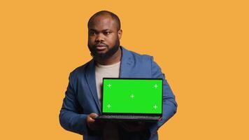 Salesman holding isolated screen laptop, doing recommendation, talking with audience. BIPOC person pointing towards chroma key notebook and showing thumbs up sign, studio background, camera A video