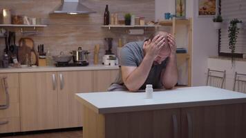 Exhausted man holding a bottle of pills sitting in the kitchen. Stressed, tired unhappy worried unwell person suffering of migraine, depression, disease and anxiety feeling ill with dizziness symptoms video