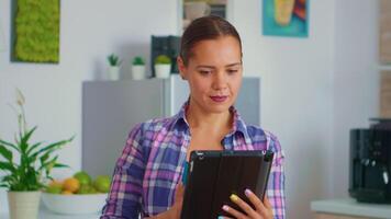 vrouw gebruik makend van tablet zittend Bij de tafel in de keuken. dame glimlachen lezing nieuws Aan tablet terwijl drinken groen thee werken van huis gebruik makend van apparaat met internet technologie typen, browsen Aan apparaatje. video