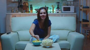 contento mujer gasto su gratis hora acecho televisión comiendo palomitas de maiz y Bebiendo jugo. emocionado divertido hogar solo dama disfrutando el noche a hogar sentado en cómodo sofá vestido en pijama. video