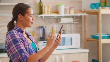 mulher usando Smartphone e bebendo verde chá, gastar livre Tempo em a cozinha. segurando telefone dispositivo com tela sensível ao toque usando Internet tecnologia rolagem, navegando, procurando em gadget durante café da manhã video