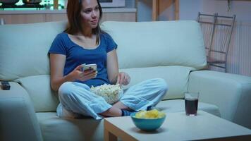 mujer desplazamiento en teléfono comiendo palomitas de maiz y acecho un película. solitario divertido contento dama lectura, escribiendo, buscando, hojeada en teléfono inteligente riendo divertido utilizando tecnología Internet relajante a noche. video