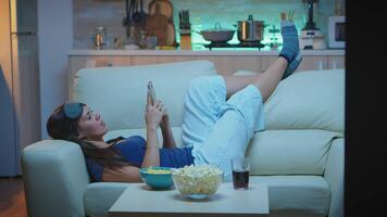 Woman with pijamas lying on sofa checking cell phone with sleep mask on forehead. Lonely happy lady relaxing using smartphone sitting on couch reading, writing, searching, browsing on mobile internet video