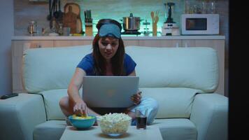 Woman with eye mask using laptop at night while watching tv and eating snacks. Happy person in pijamas sitting on sofa reading writing searching browsing on notebook using internet checking mails video