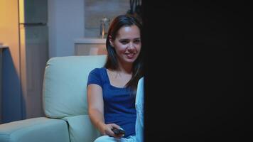 Amused lady watching Tv show in the night sitting on couch. Young happy excited home alone woman enjoying the evening sitting on comfortable sofa dressed in pajamas eating popcorn in front television video