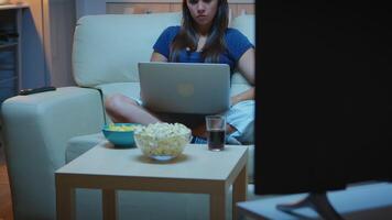 Woman freelancer in pijamas working late at night on laptop in front tv. Happy person in pijamas with eye mask sitting on sofa reading writing searching browsing on notebook using internet technology video