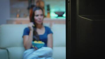 Lady eating snacks while watching tv sitting on sofa. Young happy, excited, amused, home alone lady in pajamas enjoying the evening sitting on comfortable couch eating popcorn in front of television. video