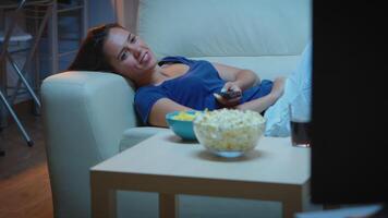 Young woman in pijamas resting lying on couch in front tv smiling. Happy, amused, lonely lady enjoying the evening sitting on comfortable sofa watching television eating popcorn and snacks video