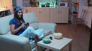 Woman sitting on sofa playing game late at night wering eye mask on forehead. Excited determined gamer using controller joysticks keypad playstation gaming and having fun winning electronic game video