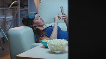 Lady with sleep mask on forehead and pijamas searching on phone lying in sofa. Happy woman sitting on couch reading, searching, browsing on smartphone using mobile internet video