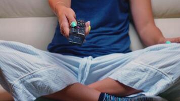 proche en haut coup de manette dans femme main tandis que séance sur canapé. éloigné contrôle dans le mains de la personne montrer du doigt à la télé, pressage le bouton et en changeant canaux séance dans de face de télévision. video