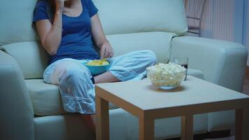 Hablando en teléfono participación meriendas cuenco y comiendo meriendas en frente de televisor. feliz, relajado, solitario dama en pijama disfrutando el noche sentado en cómodo sofá en frente de televisión utilizando teléfono inteligente video