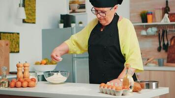 Baker spreading flour on wooden table at home in modern kitchen wearing apron and bonete. Happy elderly chef with uniform sprinkling, sieving sifting raw ingredients by hand baking homemade pizza video