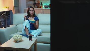 Young woman in pajamas staying up late at night eating snacks and watching tv. Bored, home alone late at night lady relaxing watching tv lying on comfortable couch holding bowl and smiling. video