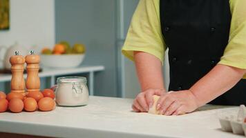Baker preparing fresh breads and cakes at home in modern kitchen. Retired elderly baker with bonete mixing ingredients with wheat flour kneading dough for baking homemade cake and bread video