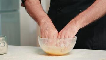 Senior chefe de cozinha aspersão farinha e amassar massa dentro vidro tigela. fechar aposentado idosos padeiro com bonete e avental, espalhando, peneiramento peneirar cru ingredientes de mão cozimento caseiro pizza, pão. video