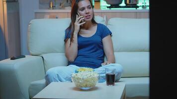 mujer en pijama hablando a teléfono acecho televisión y comiendo meriendas. feliz, relajado, solitario dama en pijama disfrutando el noche sentado en cómodo sofá en frente de televisión Hablando en teléfono inteligente video