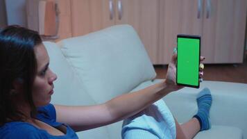 Lady reading on smartphone monitor with green screen lying on sofa in living room. Woman holding a mobile phone with mockup template chroma key isolated cell phone display using techology internet video