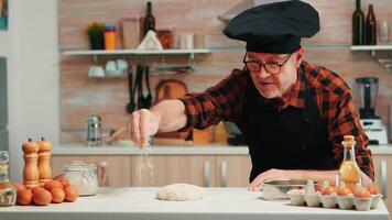 Bäckerei Mann Sieben Mehl Über Teig auf Tabelle im Zuhause Küche. im Ruhestand Alten Koch mit Bonete und Uniform streuen, Sichtung, Verbreitung rew Zutaten mit Hand Backen hausgemacht Pizza und Brot. video