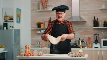 Old man with kitchen apron playing with bread dough at home smiling in front of camera. Retired elderly chef forming pizza countertop on a floured surface and kneading it with hands, in modern kitchen video