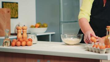 Elderly baker woman cracking eggs on glass bowl for tasty food recipe in home kitchen. Retired elderly chef with bonete mixing by hand, kneading pastry ingredients baking homemade cake. video