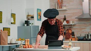 pensionato uomo a seguire culinario consigli su tavoletta, apprendimento cucinando lezione su sociale media, formatura Impasto con di legno rotolamento spillo. nonno con bonete e grembiule utilizzando il computer portatile preparazione casa torte video