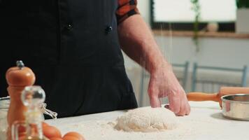 Baker adding flour on the pile by hand preparing bread dough. Close up of retired elderly chef with bonete and uniform sprinkling, sieving spreading rew ingredients baking homemade pizza and cackes. video