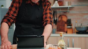 Senior homem com bonete e cozinha avental usando tábua preparando casa bolos para sobrinhos. aposentado homem Segue culinária adendo em computador portátil, Aprendendo cozinhando tutorial em social meios de comunicação, formando a massa video