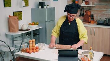 vecchio donna con bonete partecipando a culinario maestro classe su il computer portatile. pensionato capocuoco a seguire panettiere consigli su tavoletta, apprendimento cucinando lezione su sociale media, utilizzando rotolamento perno formatura il Impasto. video