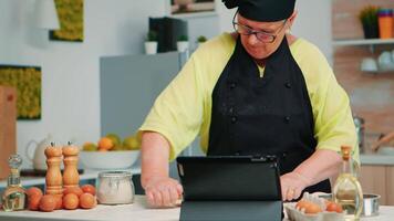 vrouw chef gebruik makend van tablet in keuken terwijl Koken pizza. gepensioneerd dame in aansluiting op culinaire advies Aan laptop, aan het leren Koken zelfstudie Aan sociaal media, gebruik makend van houten rollend pin vormen de deeg. video