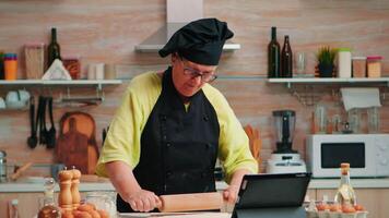 nonna ascoltando consigli per preparazione fatti in casa torte. pensionato signora a seguire culinario Podcast su tavoletta, apprendimento cucinando lezione su sociale media utilizzando di legno rotolamento perno formatura il doug video