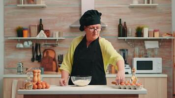 Woman baker cracking eggs into flour following traditional recipe in home kitchen. Retired elderly chef with bonete, mixing by hand, kneading in glass bowl pastry ingredients baking homemade cake video