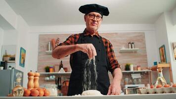 Senior man adding flour on dough by hand looking at camera smiling. Retired elderly chef with bonete and uniform sprinkling, sieving spreading rew ingredients with hand baking homemade pizza and bread video