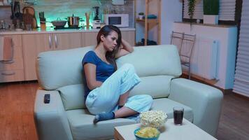 Young tired after work woman looking asleep in the evening in front of TV. Exhausted lonely sleepy lady in pajamas sleeping on sofa while watching a bored movie in living room, closing eyes at night video