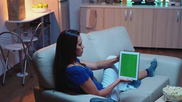 femme en portant pavé tactile avec vert écran séance sur canapé dans vivant chambre. femme au foyer à la recherche à modèle chrominance clé isolé intelligent carnet afficher en utilisant technologie l'Internet séance sur canapé. video